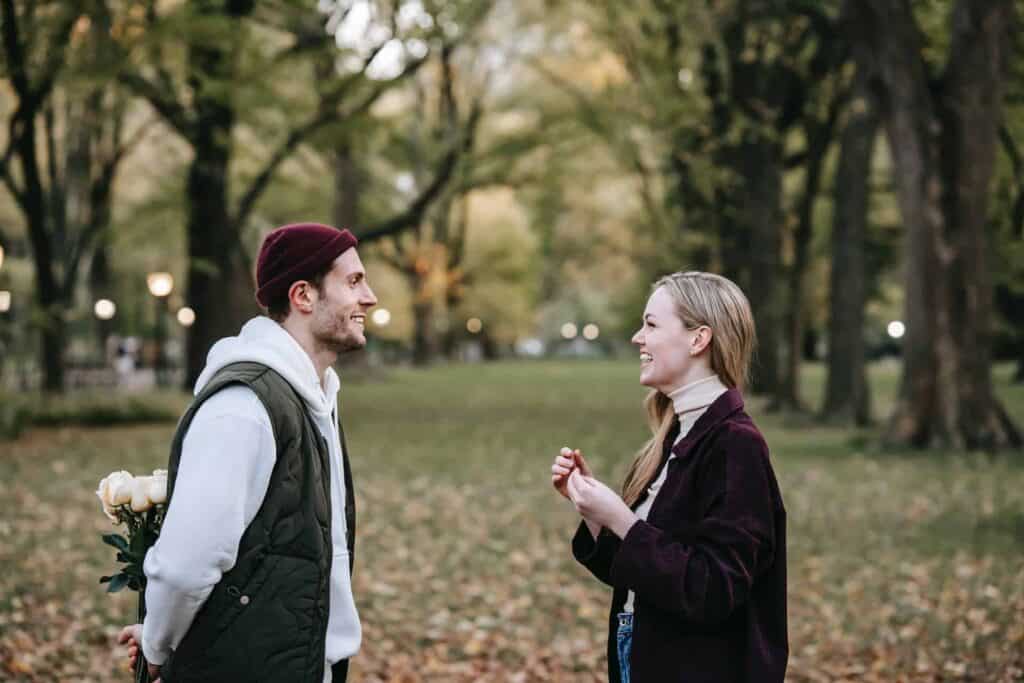 Uomo e donna che si sono appena incontrati a un appuntamento al parco, con l'uomo che tiene tra le mani dietro la schiena dei fiori